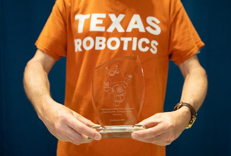A person in a Texas Robotics t-shirt holds a trophy that reads "RoboCup Standard Platform League Challenge Shield 1st Place: Eindhoven 2024"